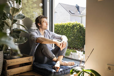 Mid adult man sitting on cushion at home on sunny day - UUF21861