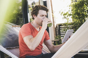 Mittelgroßer erwachsener Mann, der ein digitales Tablet benutzt und zu Hause sitzt und Saft trinkt - UUF21883
