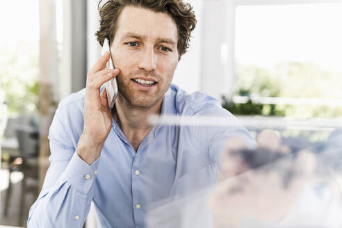 Mid adult man talking on smart phone while sitting in office - UUF21892