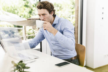 Man working on laptop while drinking coffee sitting in office - UUF21897