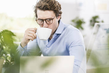 Mid adult man working on laptop while drinking coffee sitting in office - UUF21901