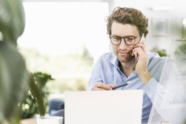 Mann spricht mit einem Smartphone, während er einen Laptop im Büro benutzt - UUF21902