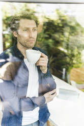 Mittlerer erwachsener Mann, der durch das Fenster schaut, während er zu Hause steht und Kaffee trinkt - UUF21906