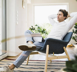 Mid adult man with hands behind head sitting on chair at home - UUF21919