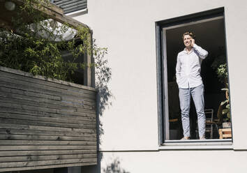 Mid adult man talking on smart phone while standing in house during sunny day - UUF21925