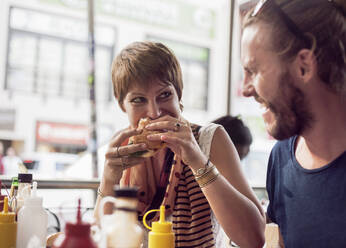 Junge Frau isst Burger, während sie mit einem erwachsenen Mann in einem Cafe sitzt - AJOF00388
