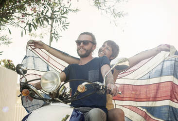 Smiling woman with arms outstretched holding national flag while sitting behind partner on motorcycle during summer - AJOF00412