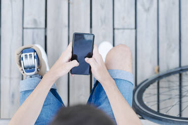 Teen male with leg prosthesis sitting on bench and browsing smartphone - CAVF89798