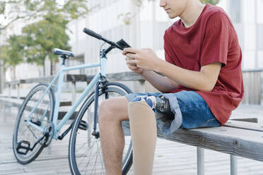 Teen male with leg prosthesis sitting on bench and browsing smartphone - CAVF89799