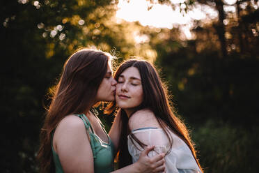Young woman kissing her girlfriend on the cheek in forest in summer - CAVF89837
