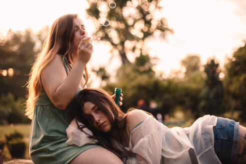 Young woman blowing bubbles while her girlfriend lying on her leg - CAVF89842