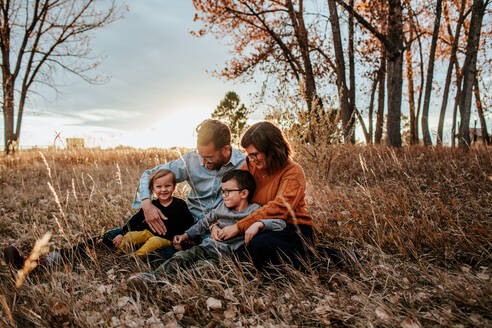 Smiling young family snuggling in a field on a fall evening - CAVF89849