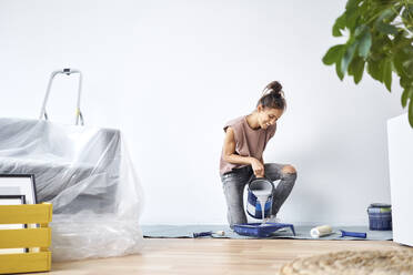 Smiling young woman pouring paint in paint tray while kneeling at home - BSZF01715