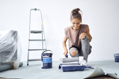 Smiling woman holding paint roller while crouching at home - BSZF01716