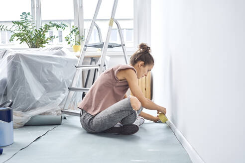 Young woman applying adhesive tape on wall while sitting at home - BSZF01720