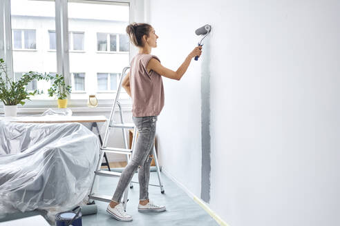 Young woman painting with paint roller on wall while standing at home - BSZF01722