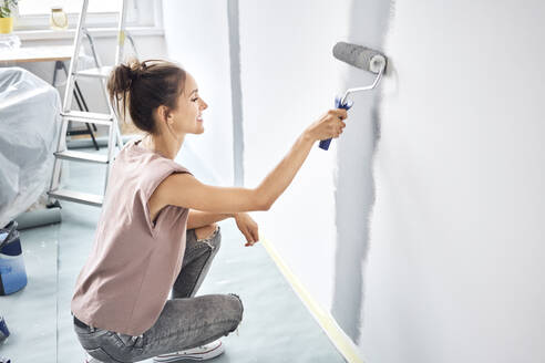 Smiling woman painting with paint roller on wall while crouching at home - BSZF01723