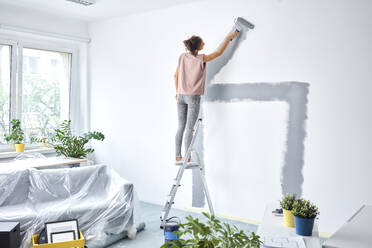 Junge Frau, die zu Hause auf einer Leiter stehend eine Wand mit einem Farbroller streicht - BSZF01724