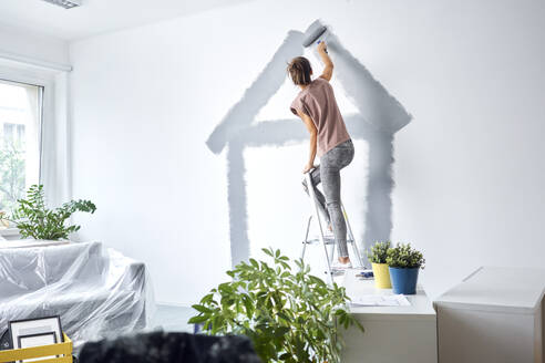 Young woman making house with paint roller on wall while standing on ladder at home - BSZF01725