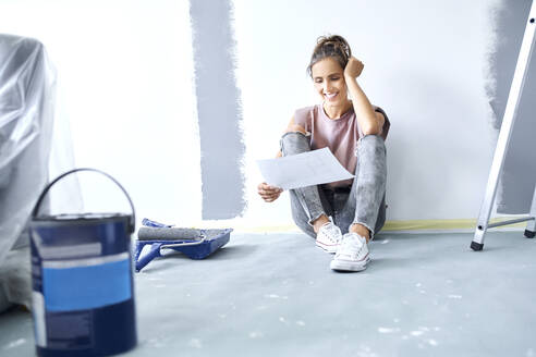 Smiling woman with head in hands holding paper while sitting on floor at home - BSZF01730