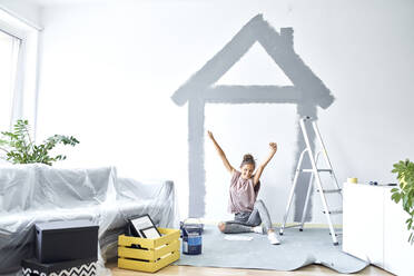 Smiling woman with hand raised sitting against wall at home - BSZF01732