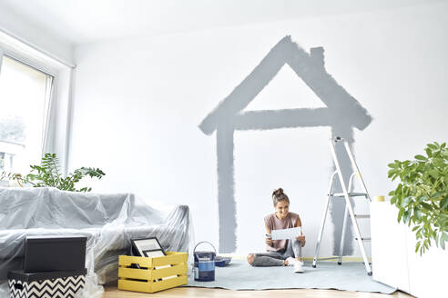 Smiling woman holding paper while sitting against wall at home - BSZF01737