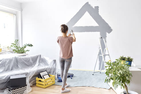 Young woman taking photo of house painted on wall through mobile phone while standing at home - BSZF01741