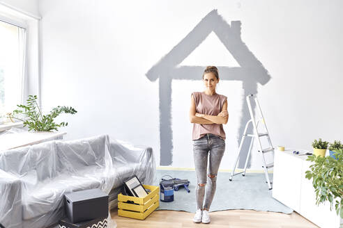 Young woman with arms crossed standing against house painted on wall at home - BSZF01742