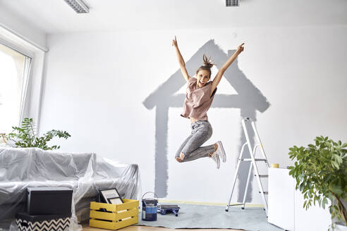 Carefree woman with hand raised jumping against wall at home - BSZF01743