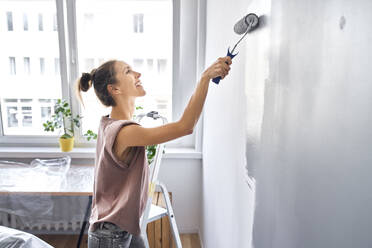Lächelnde Frau, die zu Hause stehend eine Wand mit einem Farbroller bemalt - BSZF01744