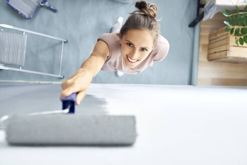 Smiling young woman painting wall with paint roller while standing at home - BSZF01745