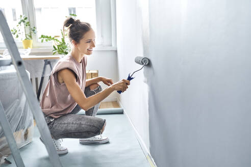 Young woman painting wall with paint roller while crouching at home - BSZF01746