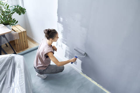 Young woman with paint roller painting wall while crouching at home stock photo