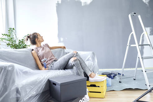 Woman with paint roller looking at wall while sitting on sofa at home - BSZF01752