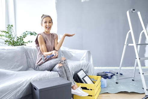 Happy woman holding paint roller while sitting on sofa at home - BSZF01753
