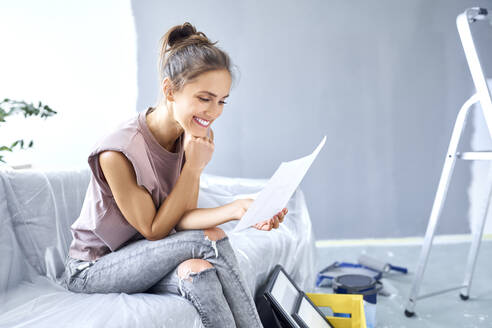 Smiling woman with head in hands reading paper while sitting on sofa at home - BSZF01754