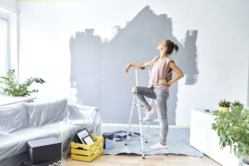 Woman with hand on hip leaning on ladder while standing against wall at home - BSZF01756