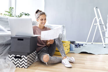 Junge Frau hält Papier, während sie zu Hause am Laptop arbeitet - BSZF01760