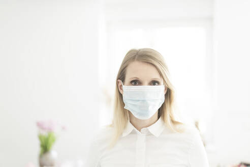 Blond woman wearing protective face mask while standing at office - CHPF00688