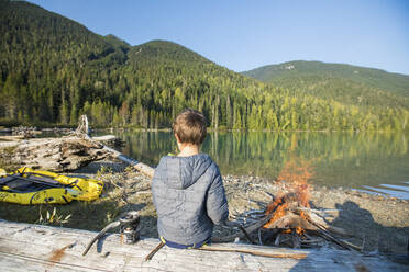 Young boy sitting on log, watching fire next to lake. - CAVF89907