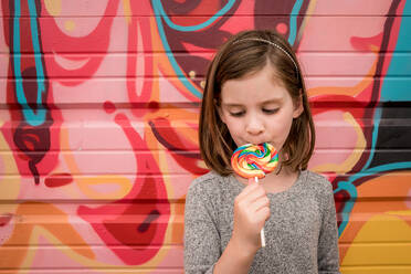 A young girls eats a huge rainbow lollipop in front of a colorful wall - CAVF89935