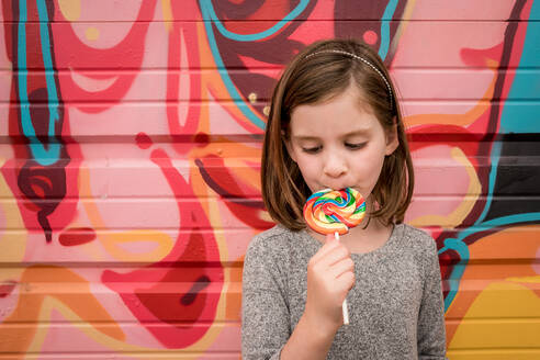 Ein junges Mädchen isst einen riesigen Regenbogen-Lolli vor einer bunten Wand - CAVF89935