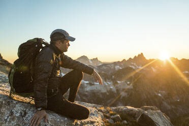 Ein fitter, aktiver Mann sitzt auf einem felsigen Bergrücken und beobachtet den Sonnenuntergang. - CAVF89991