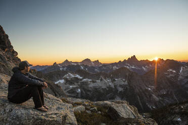 Aktiver, fitter Mann, der sich auf einem Felsen in den Bergen ausruht und den Sonnenuntergang beobachtet. - CAVF89994