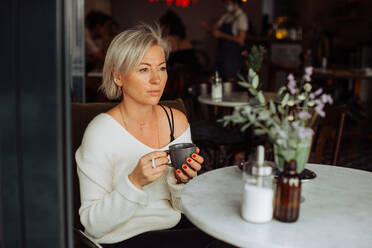 Blonde Frau, die eine Tasse Tee hält, während sie allein am Tisch im Café sitzt - CAVF90010
