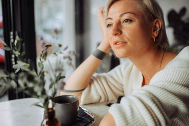 Blonde Frau sitzt am Tisch mit einer Tasse Kaffee und schaut zum Fenster - CAVF90015