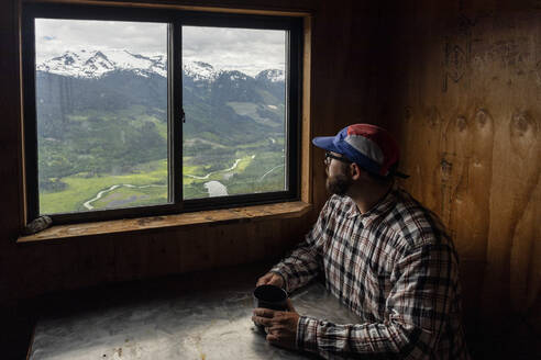 Bärtiger Mann in kariertem Hemd und Mütze sitzt am Tisch mit einem Becher Heißgetränk und schaut auf Berge, während er sich in einer gemütlichen Hütte in British Columbia, Kanada, ausruht - CAVF90017