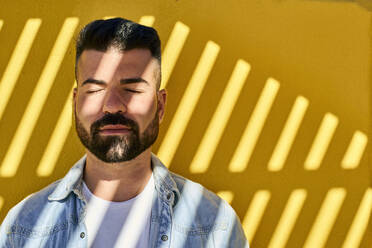 Young man with eyes closed and shadow on face standing against yellow wall outdoors - VEGF03034