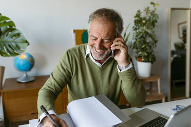 Lächelnder Mann, der in ein Buch schreibt, während er zu Hause mit seinem Handy telefoniert - VABF03620