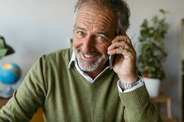 Lächelnder reifer Mann, der zu Hause sitzt und mit seinem Handy spricht - VABF03621